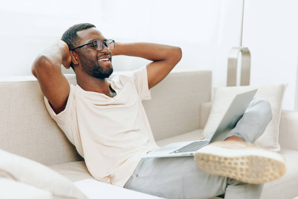 Homem Africano Americano Sorrindo Digitando no Laptop enquanto trabalhava em casa em um sofá moderno Um jovem e feliz freelancer preto, vestindo roupas casuais, senta-se confortavelmente em um sofá em sua moderna sala de estar. - Foto, Imagem
