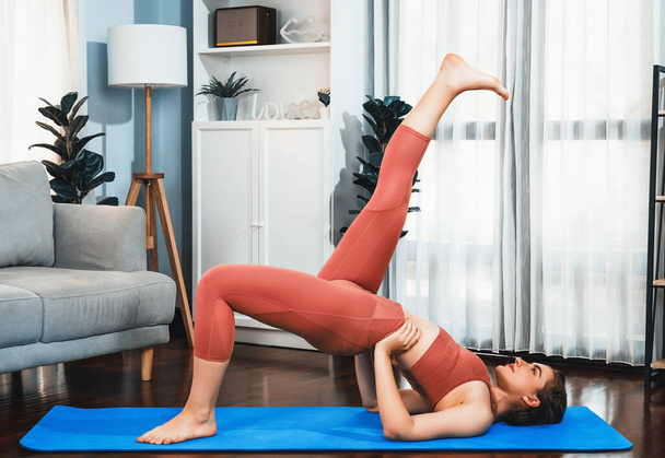 Flexible and dexterity woman in sportswear doing yoga position in meditation posture on exercising mat at home. Healthy gaiety home yoga lifestyle with peaceful mind and serenity. - Photo, Image