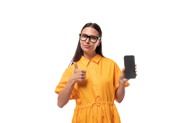 joven morena europea vestida con un vestido de verano naranja y gafas que muestran un smartphone con una maqueta. - Foto, Imagen