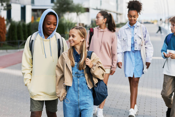 Lindos estudiantes de escuela intercultural con mochilas charlando en su camino a casa después de clases mientras caminan contra un grupo de amigos - Foto, imagen