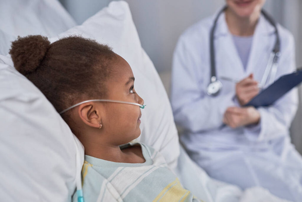 zwart klein meisje liggend in bed in ziekenhuis kamer en luisteren naar verpleegster - Foto, afbeelding