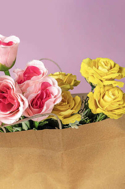 Ein Strauß künstlicher rosa und gelber Rosen in einer Papiertüte auf rosa Hintergrund - Foto, Bild