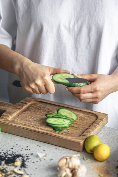 Une femme coupe un concombre frais sur une planche de bois dans la cuisine. Préparation d'une salade de légumes végétarienne. Gros plan, mains coupant des légumes. - Photo, image