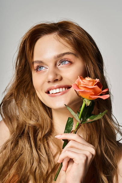 Piękna uśmiechnięta młoda kobieta z niebieskimi oczami, z brzoskwiniową różą na szarym tle - Zdjęcie, obraz