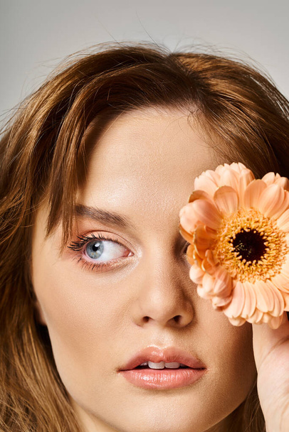 Closeup beauty shot of pretty woman with peach natural makeup and daisy near eye on grey background - Photo, Image