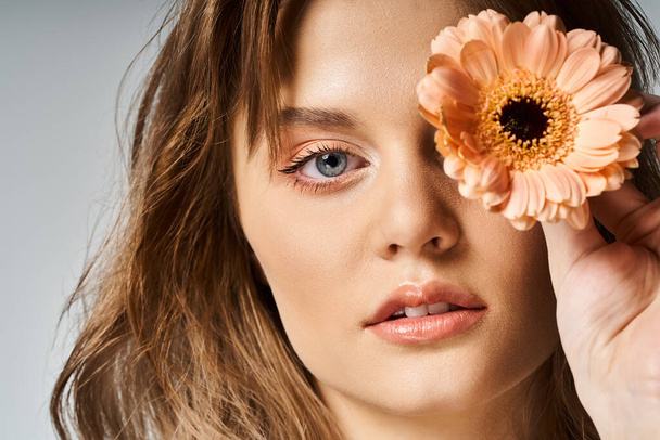 Closeup beauty shot of pretty woman with peach makeup and gerbera daisy near eye on grey background - Photo, Image