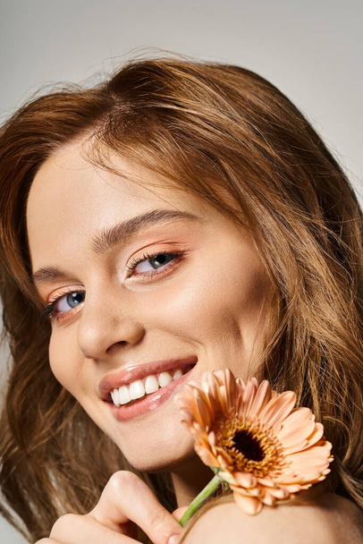 Closeup beauty shot of smiling woman with peach makeup and gerbera daisy on grey background - Photo, Image