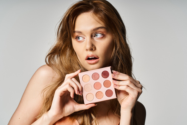 Beauty photo of surprised woman holding nude eyeshadow palette near face and looking away - Photo, Image