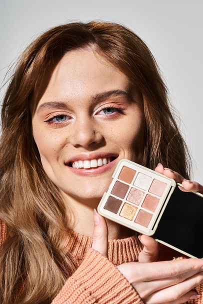 Schönheitsbild einer lächelnden Frau mit Pfirsich-Make-up-Palette am Kinn auf grauem Studiohintergrund - Foto, Bild