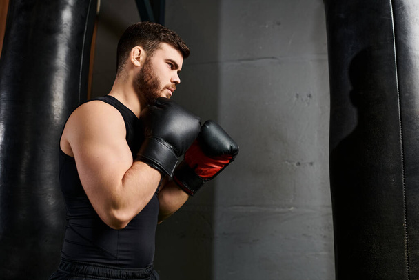 Un hombre guapo con barba que lleva una camiseta negra golpea una bolsa de boxeo en un gimnasio mientras luce vibrantes guantes rojos. - Foto, imagen