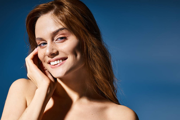 Schönheitsporträt einer lächelnden Frau, Pfirsich-Make-up und Sommersprossen, die die Wange auf blauem Hintergrund berühren - Foto, Bild