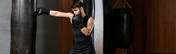 Un bell'uomo con la barba con camicia nera e guanti neri, che fa pugilato sul ring con un sacco da boxe in palestra. - Foto, immagini