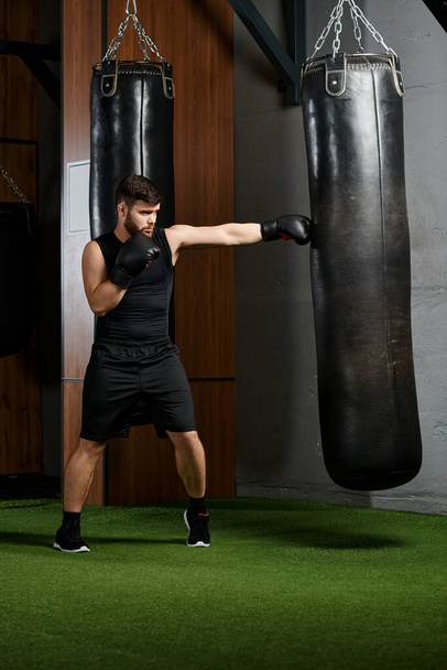Красивый бородатый мужчина в черной рубашке и шортах энергично бьет боксерскую грушу в спортзале. - Фото, изображение