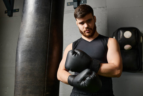 Un guapo barbudo con camisa negra y guantes de boxeo, golpeando enérgicamente una bolsa en el gimnasio. - Foto, Imagen