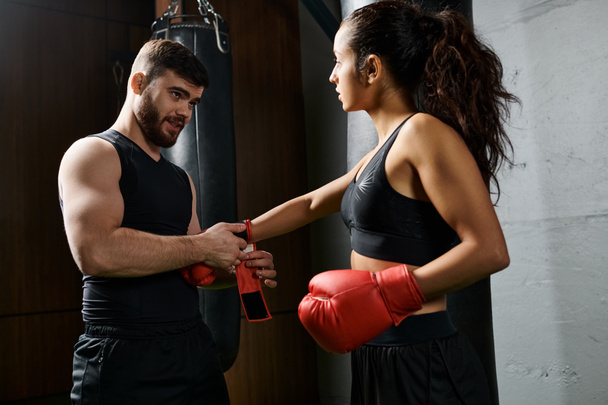 Ένας άνδρας γυμναστής στέκεται δίπλα σε μια μελαχρινή αθλήτρια με ενεργό ένδυμα καθώς φοράει γάντια πυγμαχίας και προπονείται σε ένα γυμναστήριο. - Φωτογραφία, εικόνα