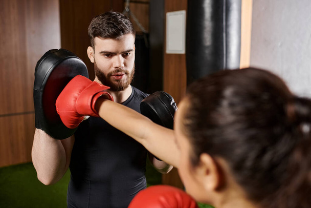 Een mannelijke trainer assisteert een brunette sportvrouw, beide gekleed in actieve kleding, als ze deelnemen aan een bokssessie in een sportschool. - Foto, afbeelding