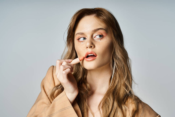 Mujer atractiva con joyas de la cara en chaqueta beige, aplicación de lápiz labial y mirando hacia otro lado, pelusa de melocotón - Foto, imagen