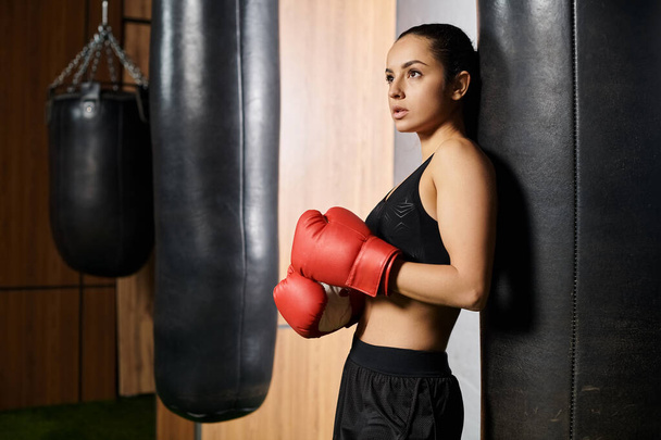 Een zelfverzekerde brunette sportvrouw in actieve kleding staat naast een bokszak met rode bokshandschoenen, klaar voor een intensieve training. - Foto, afbeelding
