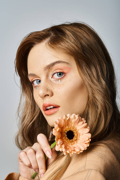 Pretty woman looking at camera and holding daisy, wearing peach makeup, face jewels - Foto, afbeelding