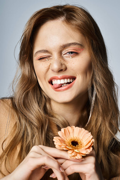 Подмигивая веселой женщине, смотрящей в камеру и держащей маргаритку, носящей персиковый макияж, украшения для лица - Фото, изображение