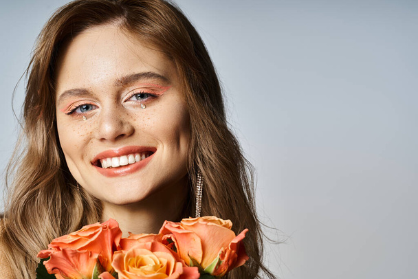 Mujer sonriente mirando a la cámara, sosteniendo rosas y usando maquillaje de melocotón desnudo y joyas de la cara - Foto, Imagen