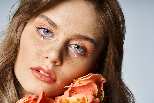 Closeup beauty shot of woman with peach makeup, face jewels and freckles, holding roses near cheek - Photo, Image