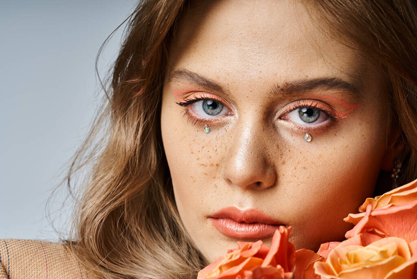 Closeup beauty shot of woman with peach makeup, face jewels and freckles, holding roses near cheek - Photo, Image