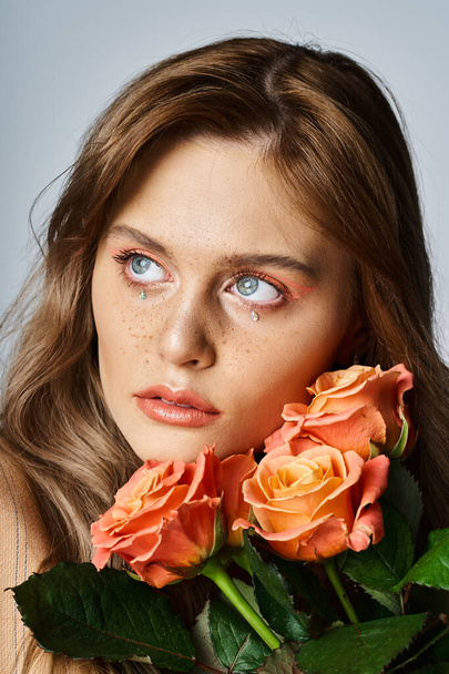 Mulher atraente com rosas perto de seu rosto e vestindo maquiagem pêssego nu e jóias rosto espumante - Foto, Imagem