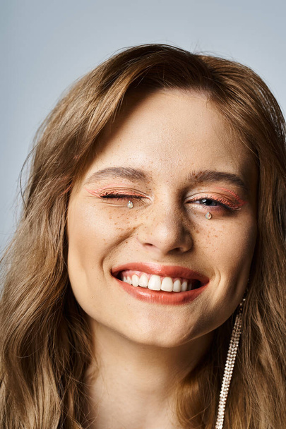 Ujęcie z bliska uśmiechniętej mrugającej kobiety z brzoskwiniowym makijażem, klejnotami do twarzy i piegami na szarym tle - Zdjęcie, obraz