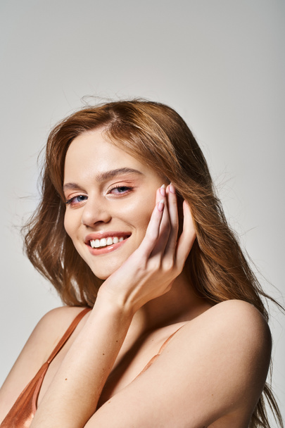 Jonge mooie vrouw met perfecte huid glimlachen op camera aanraken gezicht, op grijze studio achtergrond - Foto, afbeelding