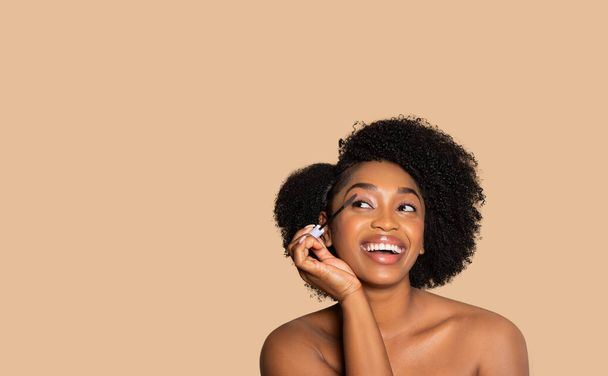 Iloinen nuori musta nainen kihara tukka soveltamalla ripsiväri, katsoen ylöspäin vapaata tilaa iloinen ilme, vastaan sohva beige studio tausta - Valokuva, kuva