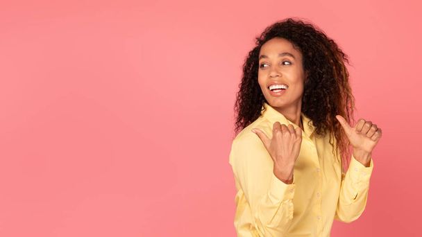 Mujer afroamericana joven con confianza apuntando hacia atrás en el espacio vacío, invitando a la atención y la curiosidad, contra el fondo rosado animado, mostrando promoción, publicidad - Foto, Imagen