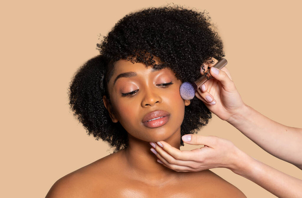 Makeup καλλιτέχνης εφαρμόζει απαλά ρουζ σε χαλαρό μαύρο μοντέλο με σγουρά μαλλιά, αποδεικνύοντας επαγγελματικές τεχνικές ομορφιάς σε ουδέτερο μπεζ φόντο - Φωτογραφία, εικόνα
