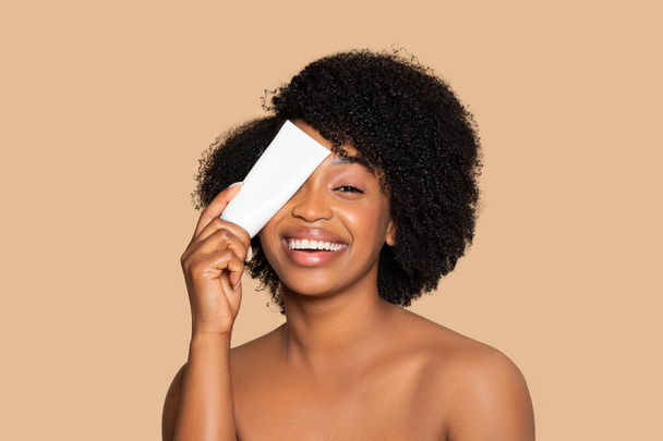 Mujer negra joven exuberante con el pelo rizado cubriendo un ojo con tubo blanco de crema, simbolizando la diversión y la simplicidad de las rutinas de cuidado de la piel, fondo beige - Foto, imagen