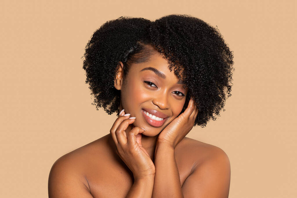 温かいベージュの背景に柔らかい笑顔を表示し,穏やかなタッチで彼女の頬を保持するキュリーな髪のコンテンツの若い黒人女性 - 写真・画像