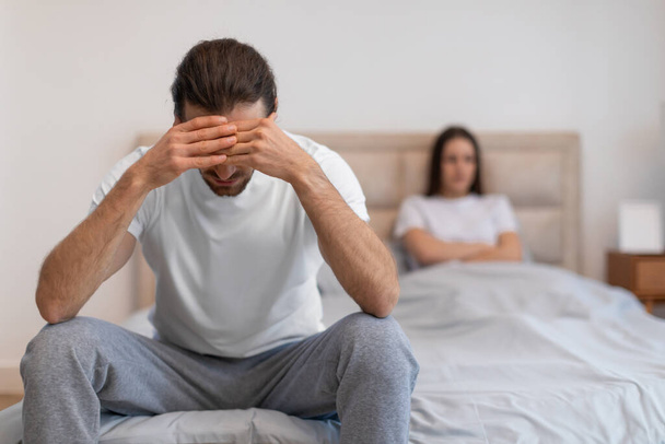 Обезумевший мужчина сидит на кровати с закрытыми руками, а обеспокоенная женщина смотрит сзади, изображая стресс в отношениях в спальне - Фото, изображение