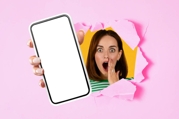 Возбужденная брюнетка демонстрирует большой экран телефона макет через отверстие в розовой стене студии. Женщина представляет пустой сенсорный экран своего мобильного гаджета рекламное предложение, приложение. Коллаж - Фото, изображение