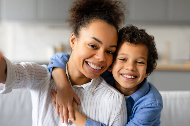 Avec un sourire joyeux, mère noire et son fils préadolescent prenant selfie, capturant moment de bonheur et de proximité dans le salon à la maison - Photo, image