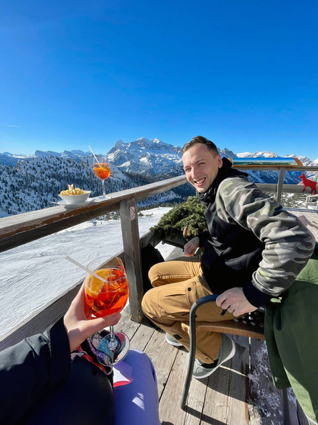 A handsome young man enjoying a break against the backdrop of mountains after snowboarding, sipping on an apres ski drink. Alpine scenery, mountain view. - Photo, Image