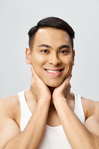 Un hombre asiático guapo con una sonrisa alegre, con las manos apoyadas en su barbilla en un estudio gris. - Foto, imagen