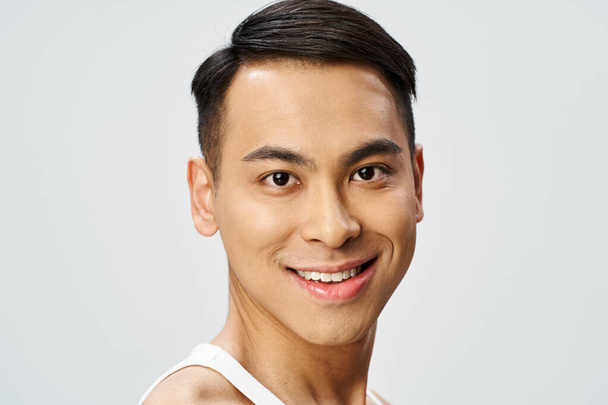 Um homem asiático bonito em um top de tanque branco sorrindo calorosamente para a câmera em um estúdio cinza. - Foto, Imagem