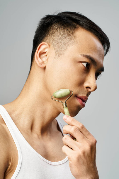 Hombre asiático guapo cuidadosamente usando rodillo de jade en un moderno estudio gris. - Foto, imagen