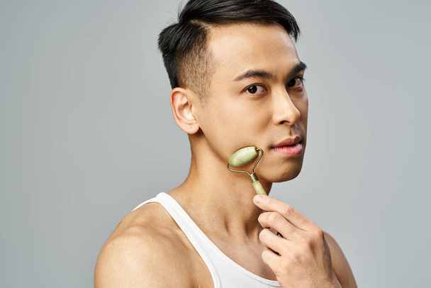 Aziatische man met behulp van jade roller, gericht op verzorging en self-care routine in een grijze studio setting. - Foto, afbeelding