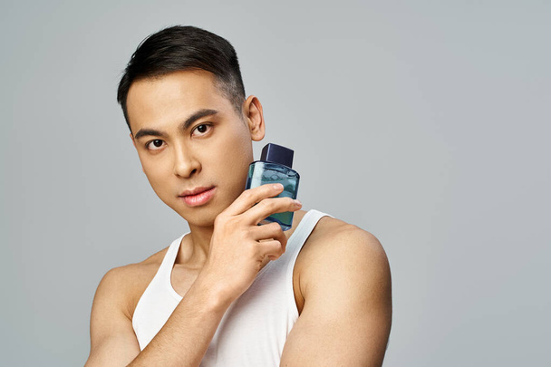 Um homem asiático bonito em um top de tanque segura uma garrafa de perfume em um estúdio cinza. - Foto, Imagem