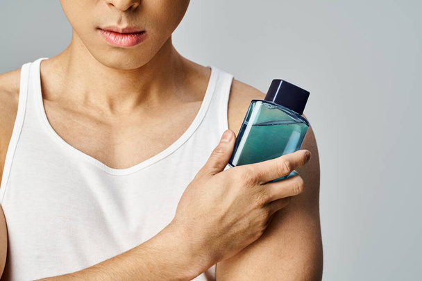 Ein zierlicher asiatischer Mann in einem Tanktop hält eine Flasche Parfüm in raffinierter Weise in einem grauen Studio-Ambiente. - Foto, Bild