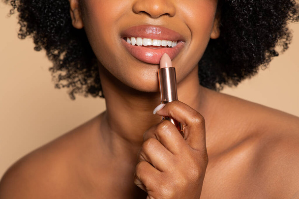 Primer plano de las mujeres negras sonríen mientras aplica lápiz labial desnudo, capturando el momento de mejora de la belleza contra el fondo beige complementario - Foto, imagen
