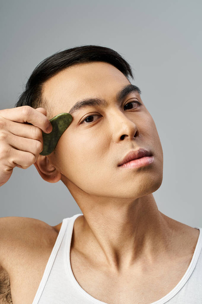 Hombre asiático guapo en un estudio gris sostiene un gua sha verde en estudio gris - Foto, imagen