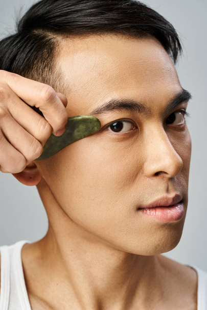 A handsome Asian man in a grey studio holds a green gua sha in his hand on grey - Photo, Image