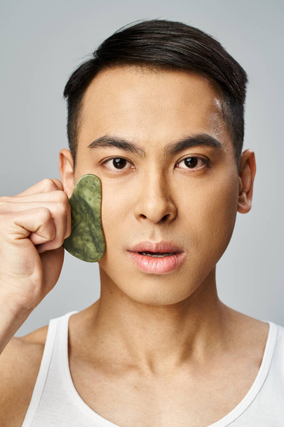 Aziatisch man met behulp van groene gua sha op zijn gezicht in een grijze studio instelling. - Foto, afbeelding