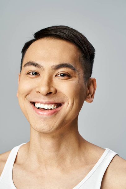 Um homem asiático bonito está irradiando com um sorriso, exalando calor e felicidade em um estúdio cinza enquanto usa produtos de cuidados com a pele. - Foto, Imagem